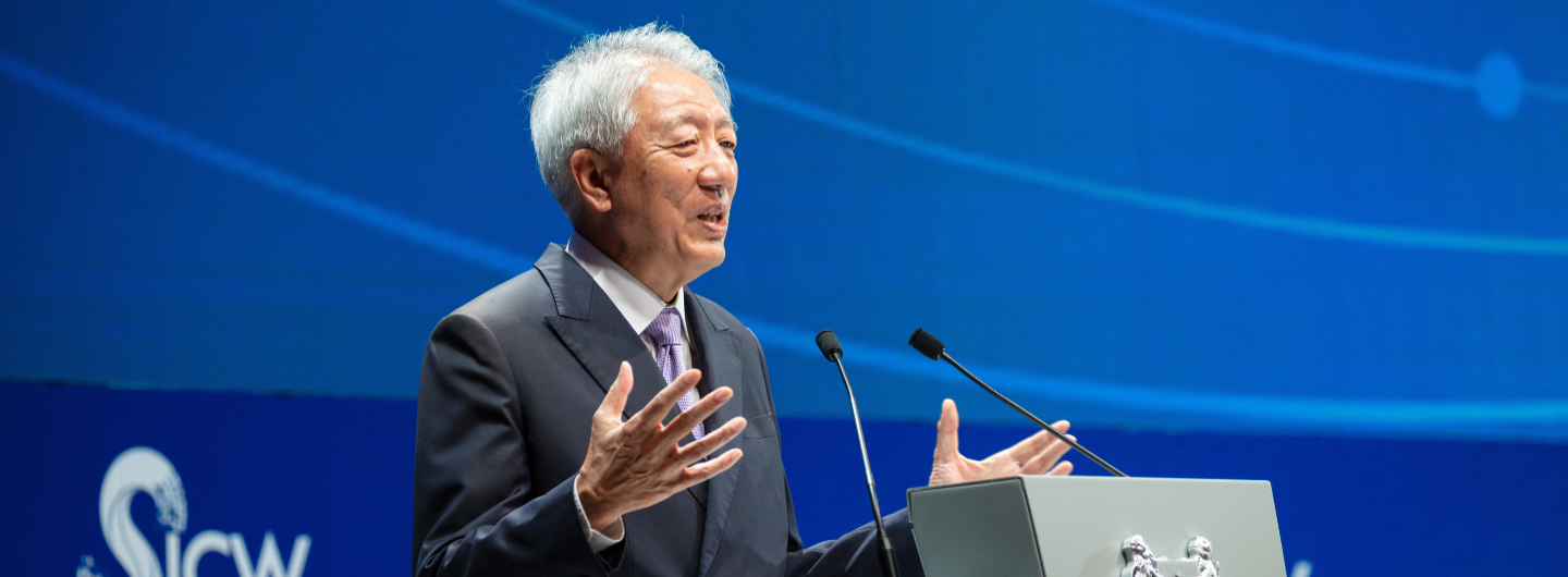 Asian man in suit at podium with mics, gesturing, blue backdrop with "SIEW" logo.
